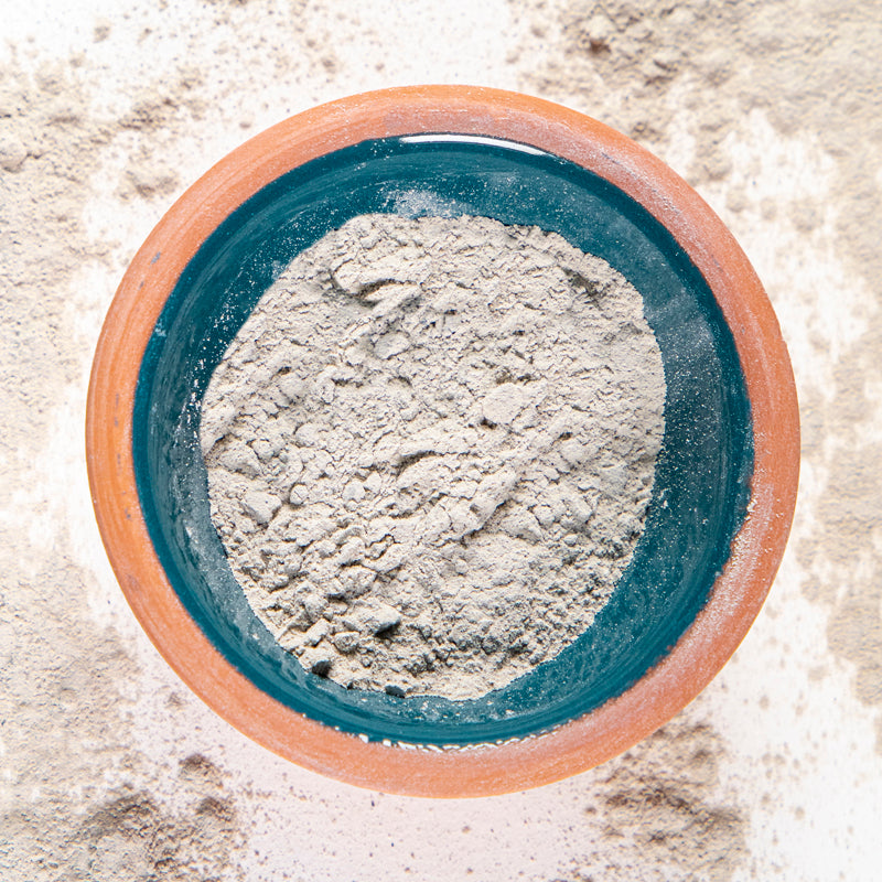 Betonite Clay in blue clay bowl with white background and herb surrounding.